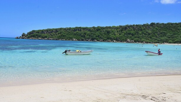 Une plage avec deux bateaux ancrés près du rivage, une plage de sable et une colline boisée en arrière-plan.