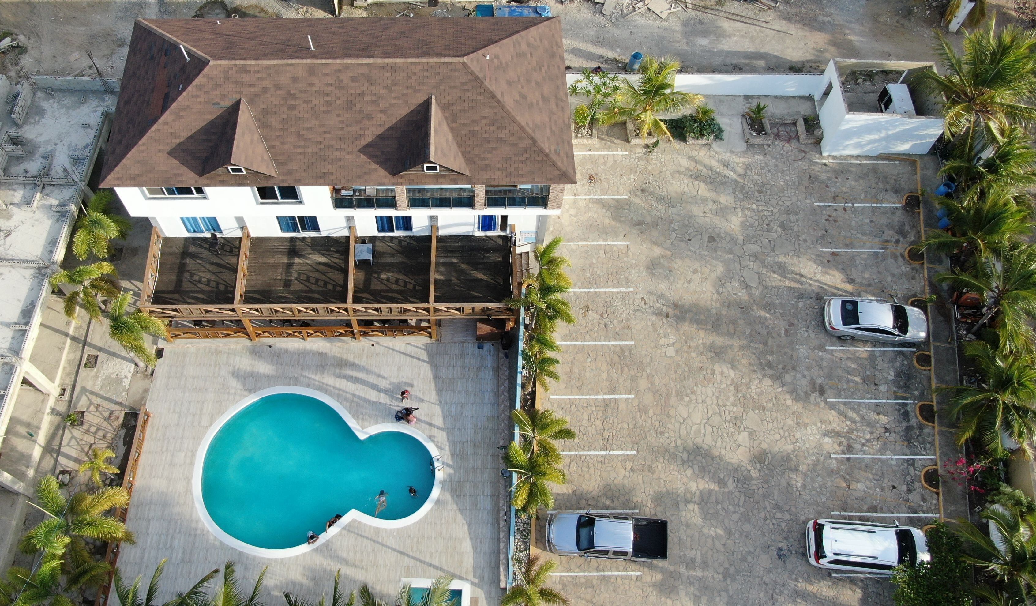 Vista aérea de un hotel con piscina, un área exterior cubierta y un estacionamiento con varios autos.