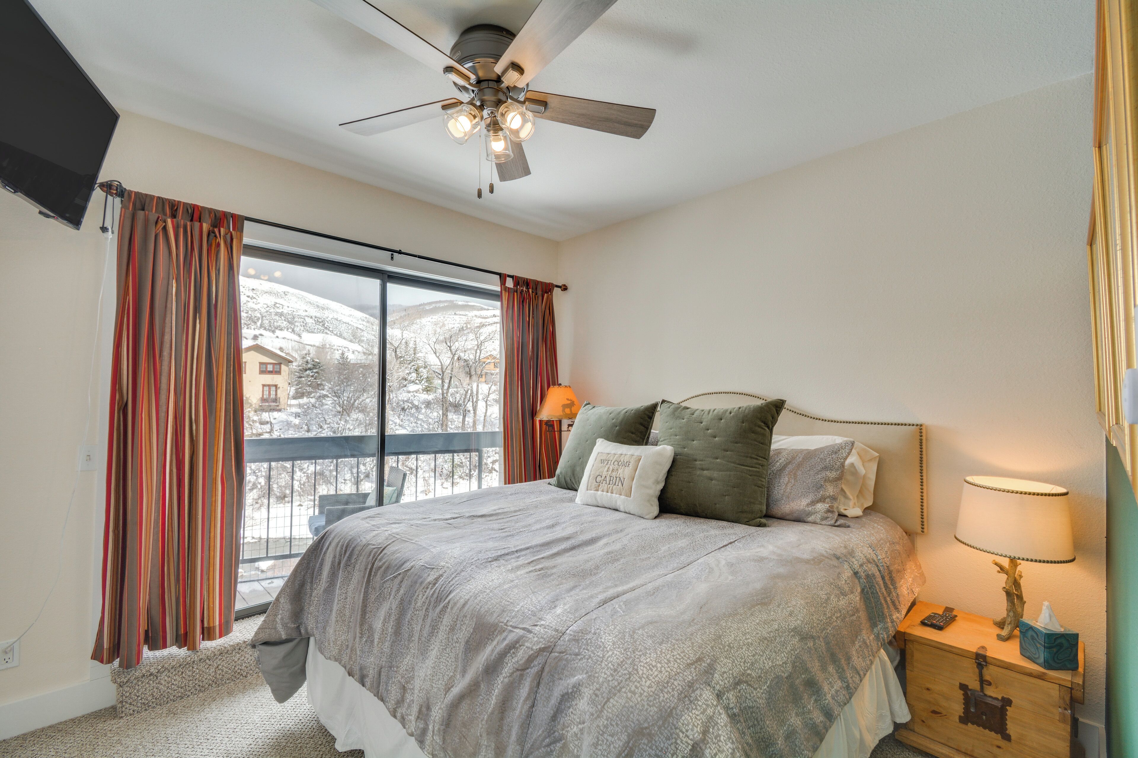 A bedroom with a large bed, a ceiling fan, a nightstand with a lamp, and a view of a snowy landscape through the window.