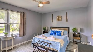 Un dormitorio con cama, ventilador de techo, ventana con cortinas, una mesita con una planta y una mesita de noche con lámpara.