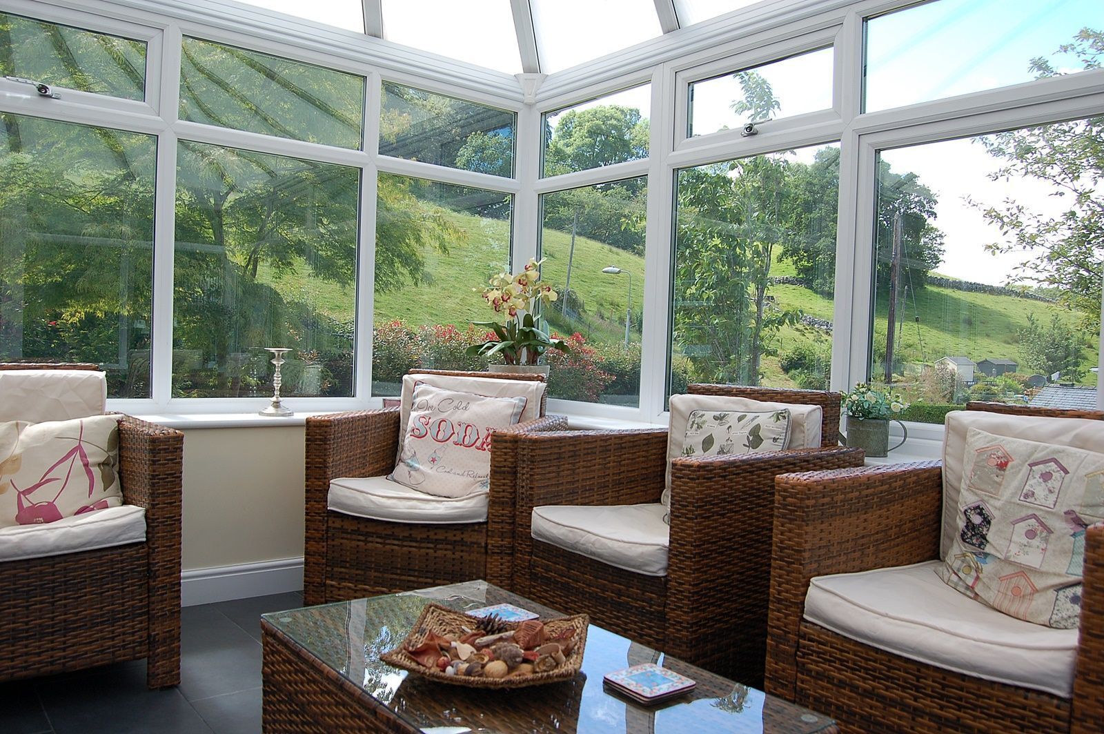 A conservatory with wicker furniture and a glass table, overlooking a green landscape.