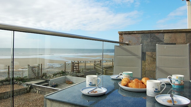 En balkon med udsigt over stranden, et bord med mad og kopper, samt en stol.