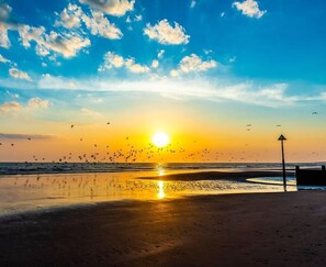 A beach scene with the sun setting over the horizon, birds flying, and a lifeguard stand in the distance. - Ty Traeth Beach House (Tywyn)