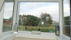 A room with a large window overlooking a fenced garden and a distant hill. - Idris View Family Holiday Home (Tywyn)