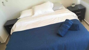 Un lit bien fait, recouvert d’une couvre-lit bleue et agrémenté d’oreillers blancs, une petite table de chevet avec une lampe, et une table de nuit noire sur laquelle se trouvent un téléphone et un récipient.