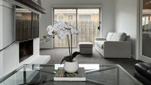 A modern living room with a glass coffee table, a white sofa, a white armchair, a fireplace, and a large window with a view of a wooden fence. - Beach Rd | Kids & Pet Friendly | Free Parking (Mordialloc)