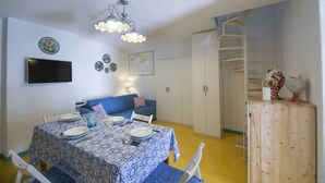 Un comedor con una mesa preparada para cuatro personas, un sofá azul, un televisor montado en la pared y un mueble de madera con objetos decorativos.