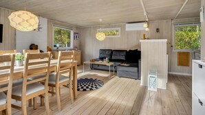 Ett mysigt och väl upplyst rum med ett matbord i trä, stolar, en soffa och ett soffbord. Det finns två fönster som släpper in naturligt ljus och ger utsikt över grönområden utanför.