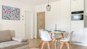 Un soggiorno moderno con un divano beige, un tavolo da pranzo con sedie bianche, una cucina con forno integrato e un quadro astratto colorato appeso al muro.