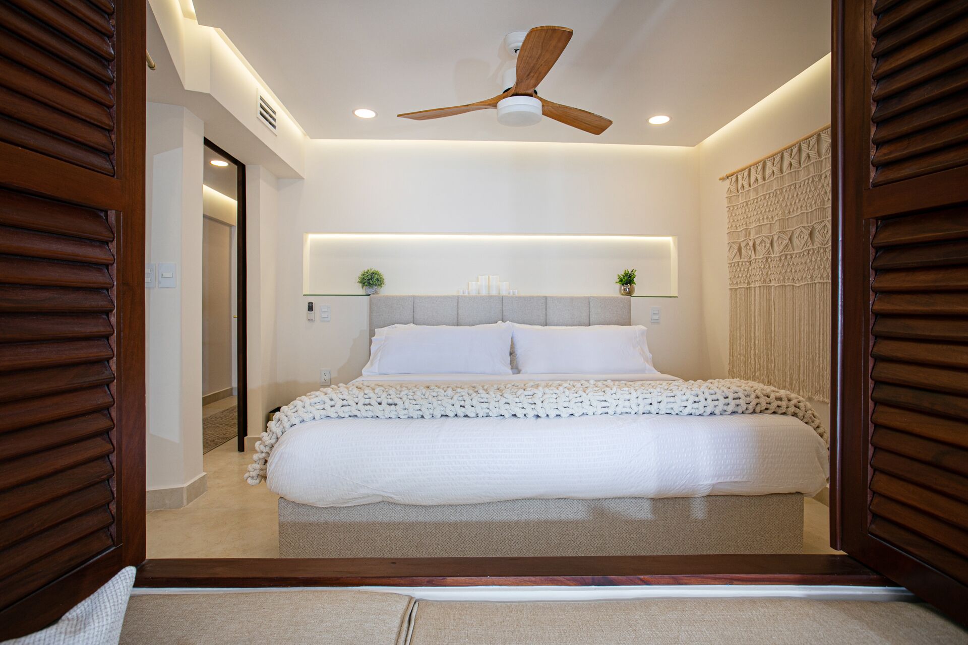 Ein modernes Schlafzimmer mit einem großen Bett, einem Deckenventilator, eingelassener Beleuchtung und einer dekorativen Wandverkleidung.