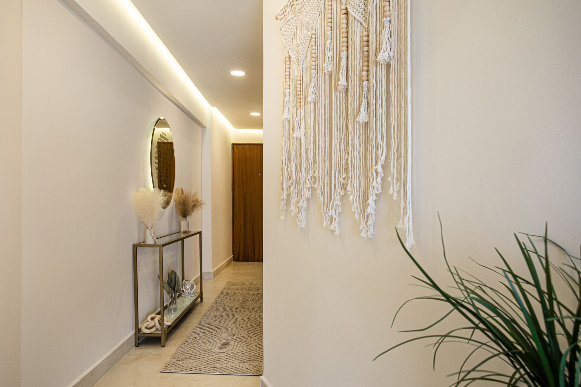 Un couloir avec une porte en bois, une console ornée d’objets décoratifs, une décoration murale en macramé et une plante en pot.