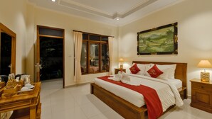 A well-lit bedroom with a large bed, wooden headboard, and a painting above it. There is a wooden table with items on it and a window with curtains. - Gora House Bali (Ubud)