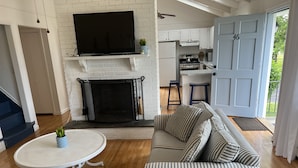 A living room with a fireplace, a television, a striped sofa, and a dining area with a kitchen in the background. - Beach Cottage (Westerly)