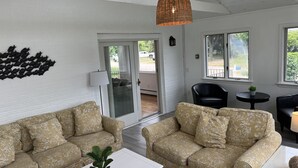 A spacious living room with two patterned sofas, a white coffee table, a hanging light fixture, and a view through a glass door to the outside. - Beach Cottage (Westerly)