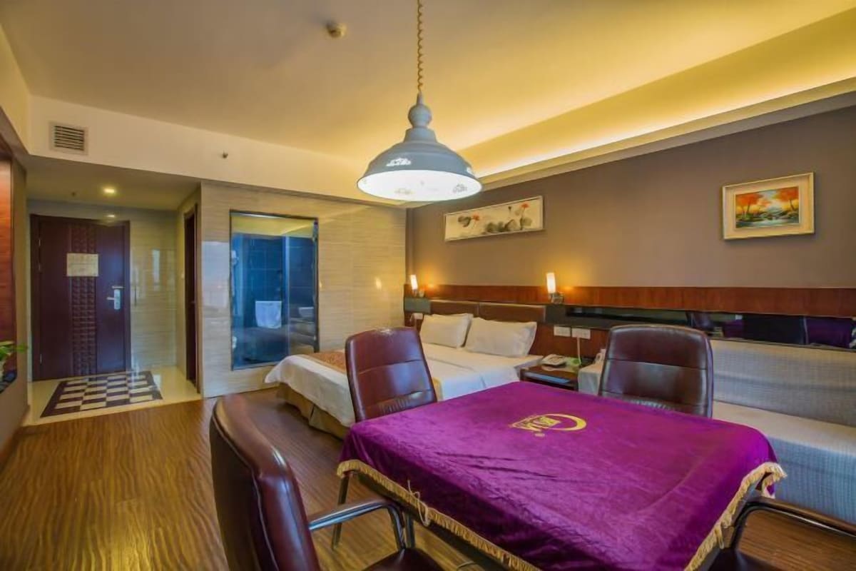 A hotel room with a bed, two chairs, a table with a purple tablecloth, a hanging light fixture, and a painting on the wall.