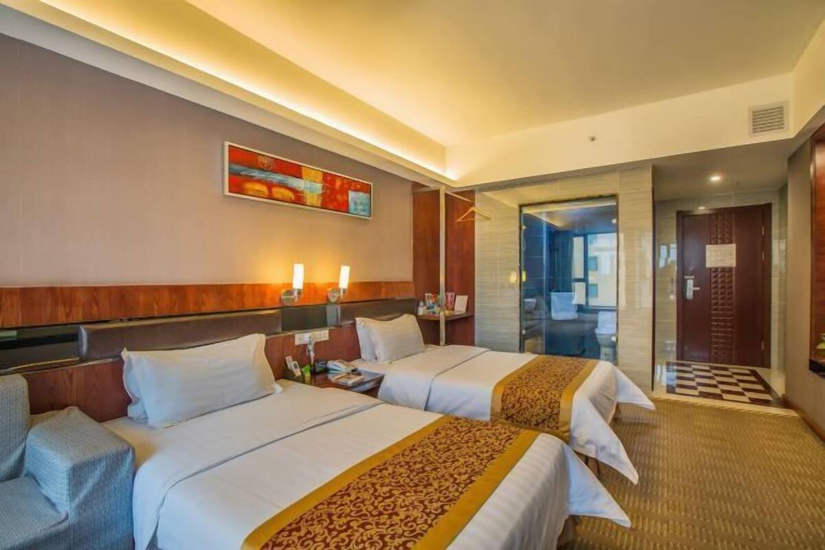 Een hotelkamer met twee bedden, een bureau, een stoel, een schilderij aan de muur en een badkamer die door een glazen deur zichtbaar is.