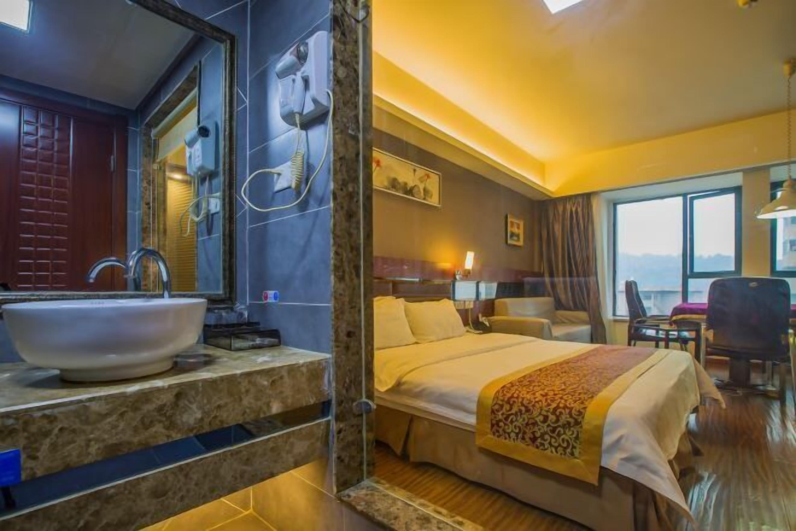 A hotel room with a large bathroom sink, a bed with a patterned bedspread, a desk with a chair, and a window with a view.