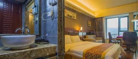 A hotel room with a large bathroom sink, a bed with a patterned bedspread, a desk with a chair, and a window with a view.