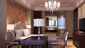 Una habitación de hotel con cama, escritorio con lámpara, mesa de comedor con sillas, televisor y candelabro.