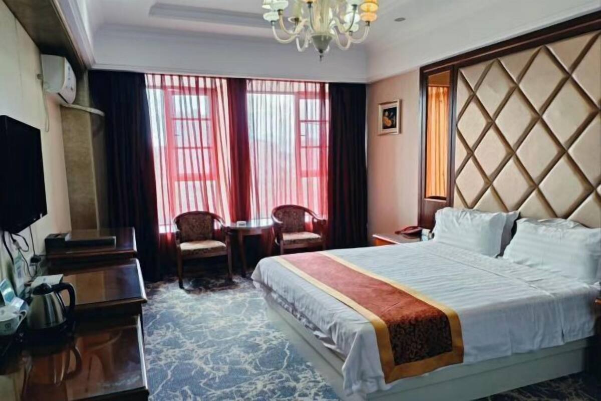 Hotelkamer met een groot bed, een bureau met waterkoker, twee stoelen, een kroonluchter en een raam met gordijnen.