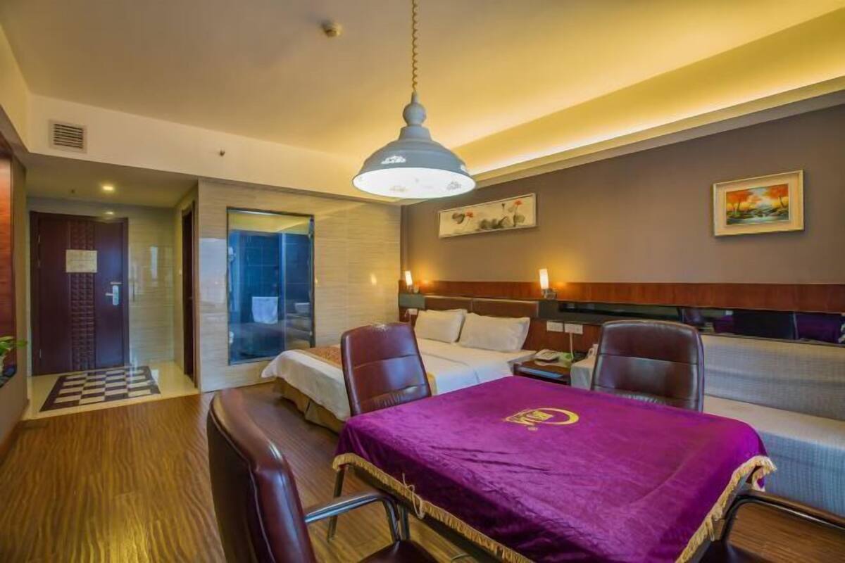 Een hotelkamer met een bed, een bureau met een paarse tafelkleed, twee stoelen, een groot raam en een schilderij aan de muur.