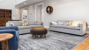 A spacious living room with two grey sofas, a blue armchair, and a round ottoman. A wooden cabinet and a kitchen area are visible in the background. - Bright & Modern 5 BR Wintergreen Home; Minutes from Hiking, Skiing, and Wineries (Wintergreen)