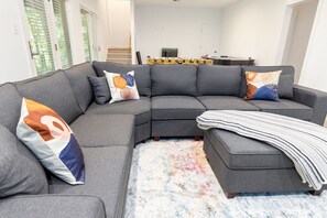 Un canapé d’angle gris agrémenté de coussins colorés, d’un plaid à rayures et d’un tapis à motifs, dans un salon moderne avec une cuisine en arrière-plan.