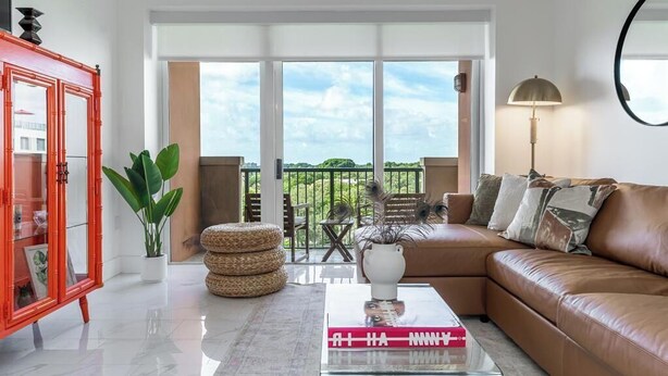 A modern living room with a brown leather sofa, a glass coffee table with a book, a red cabinet, and a large window with a view of greenery. - Coconut Grove Retreat, bayviews, 1 park incl, wkspace, Smart TV, Pool/hottub" (Coconut Grove)