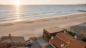 A beach view with a building in the foreground, a clear sky, and the sun setting over the ocean.
