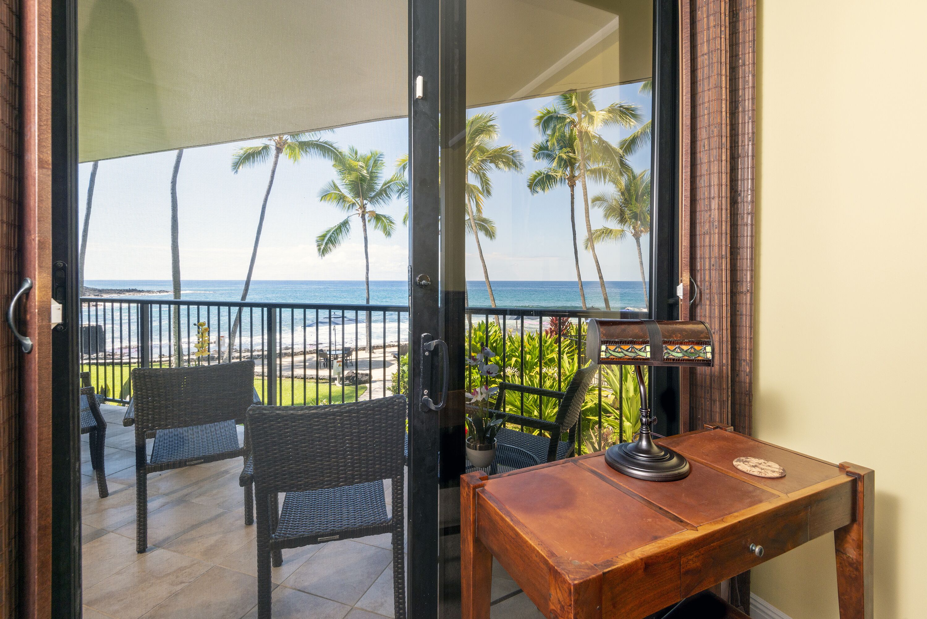 A balcony with wicker chairs, a wooden table, and a view of the ocean with palm trees.