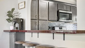 A kitchen with a stainless steel microwave, a coffee machine, and a tiled backsplash. There are three stools and a potted plant. - ❤ 5 Min Walk to Beach Fast Wi-Fi Arcade Games ❤ (Muskegon)