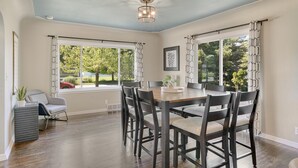 A dining area with a wooden table and chairs, large windows with curtains, and a ceiling light fixture. - ❤ 5 Min Walk to Beach Fast Wi-Fi Arcade Games ❤ (Muskegon)