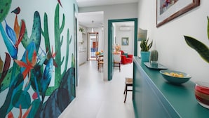Un couloir orné d’une fresque colorée, un coin repas avec une chaise rouge et une cuisine avec un cactus.