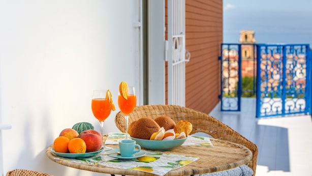 Une table en osier, sur laquelle se trouvent un saladier rempli de viennoiseries, une assiette de fruits et deux verres de jus d’orange, est placée sur un balcon offrant une vue sur la mer.