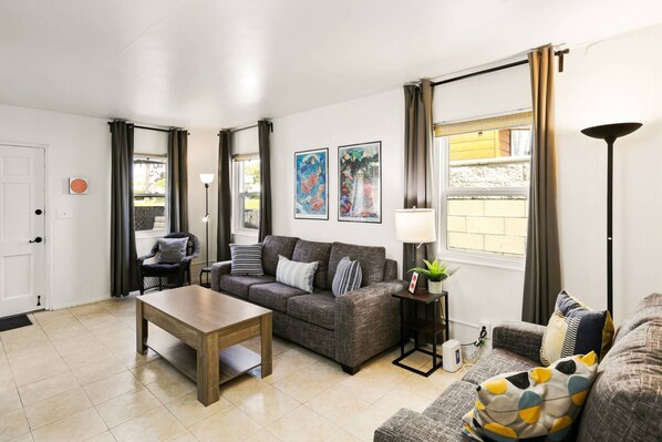 A living room with a sofa, a coffee table, and a lamp. There are two windows with curtains, a door, and a wall with two framed pictures.