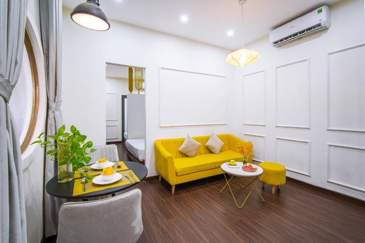 Una habitación moderna con un sofá amarillo, una pequeña mesa redonda y un taburete amarillo. Hay una planta en maceta sobre la mesa y una lámpara colgante.