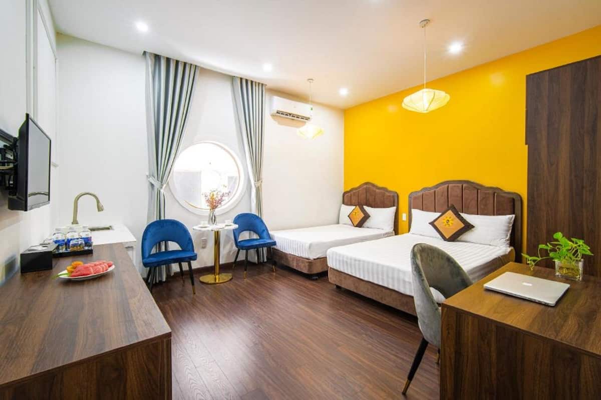 Habitación de hotel con dos camas individuales, un escritorio con una computadora portátil, una silla, una mesita con un florero y una ventana con cortinas.