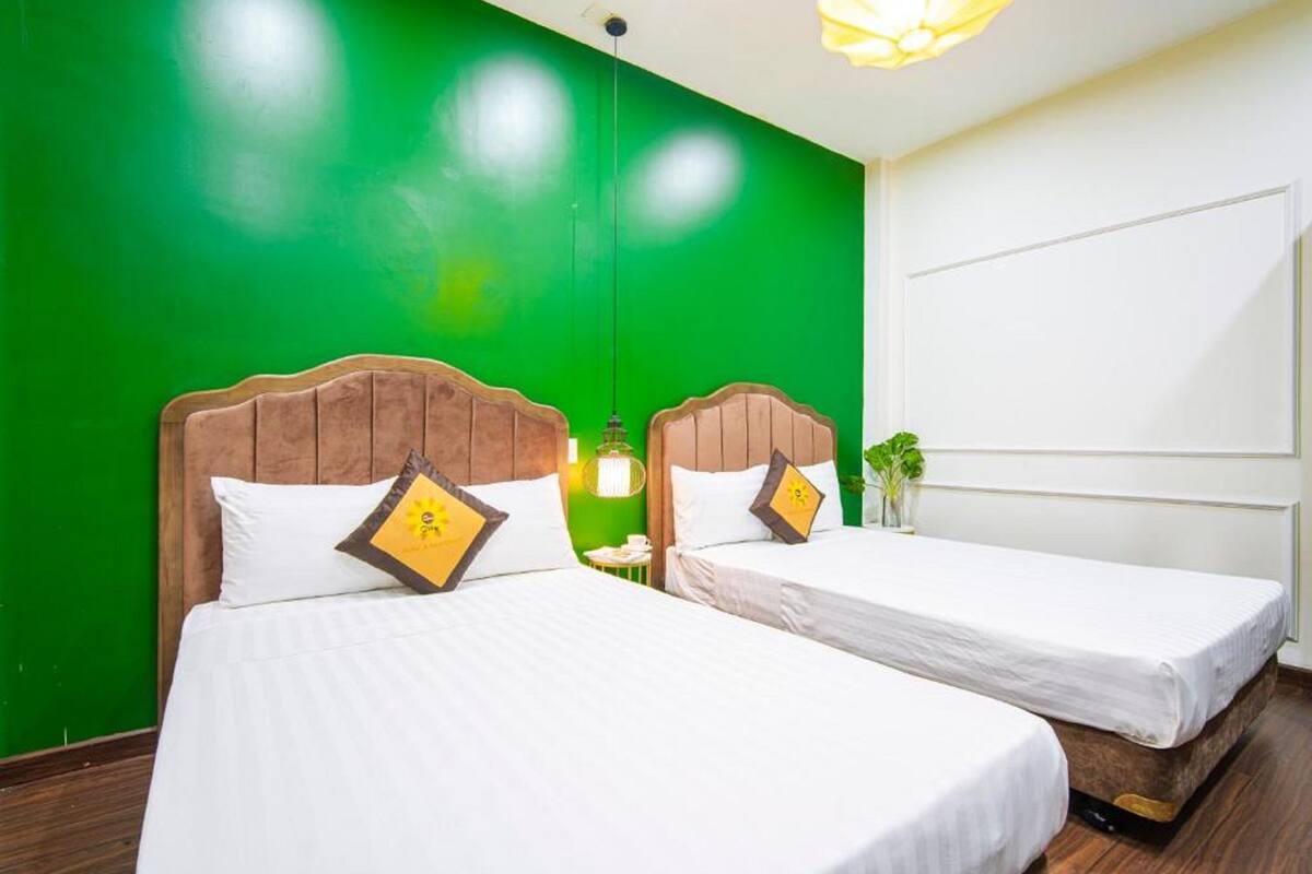 Habitación de hotel con dos camas, paredes verdes, cabeceras de madera y una mesita con lámpara.
