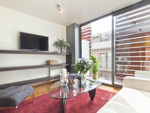 A modern living room with a glass coffee table, a flat-screen TV mounted on the wall, a grey bench, and a red rug.