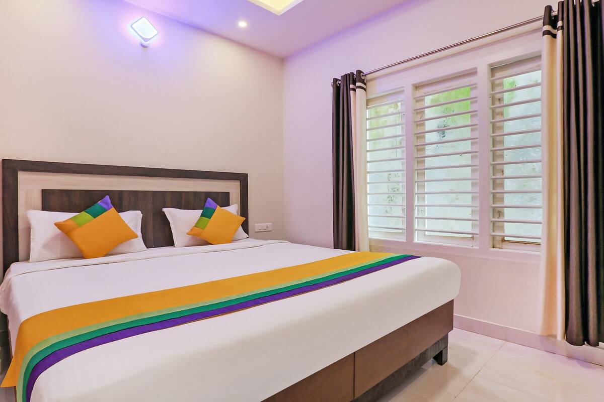 A neatly made bed with a white headboard and colorful pillows, a window with blinds, and a bedside table with a lamp.