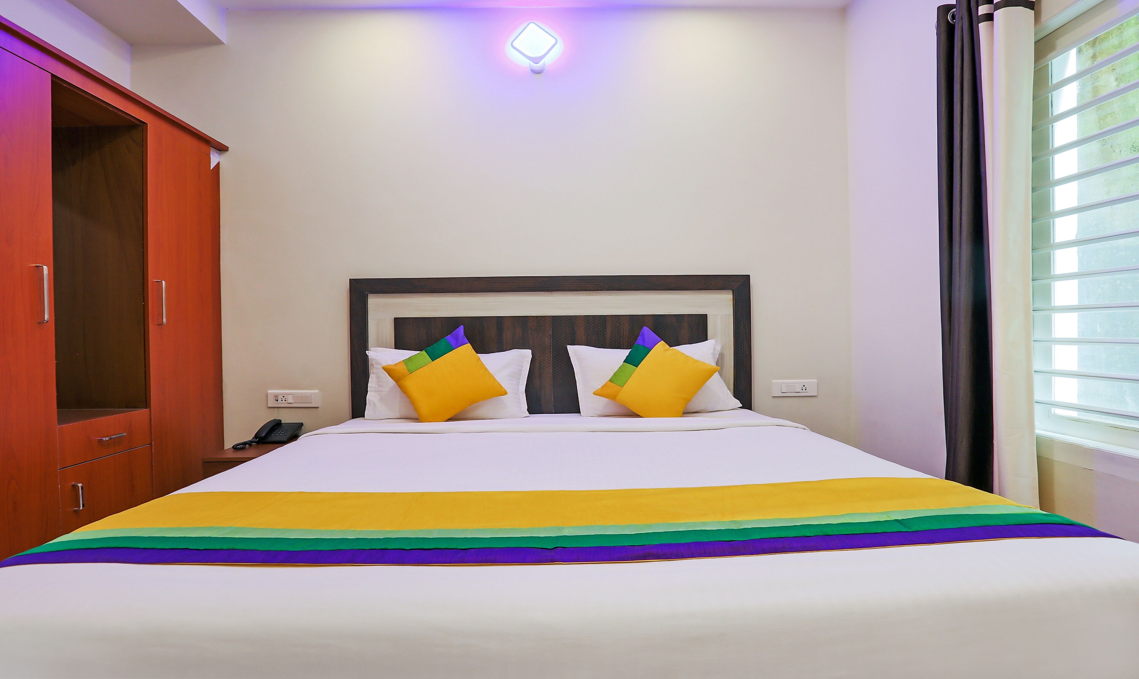 A hotel room with a bed, a wardrobe, a bedside table with a phone, and a window with curtains.