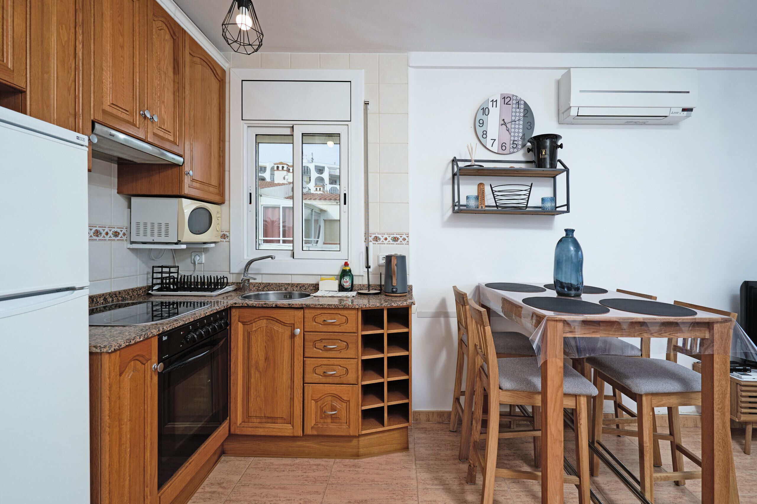 A kitchen with wooden cabinets, a microwave, a stove, and a sink. There is a dining table with chairs and a clock on the wall.