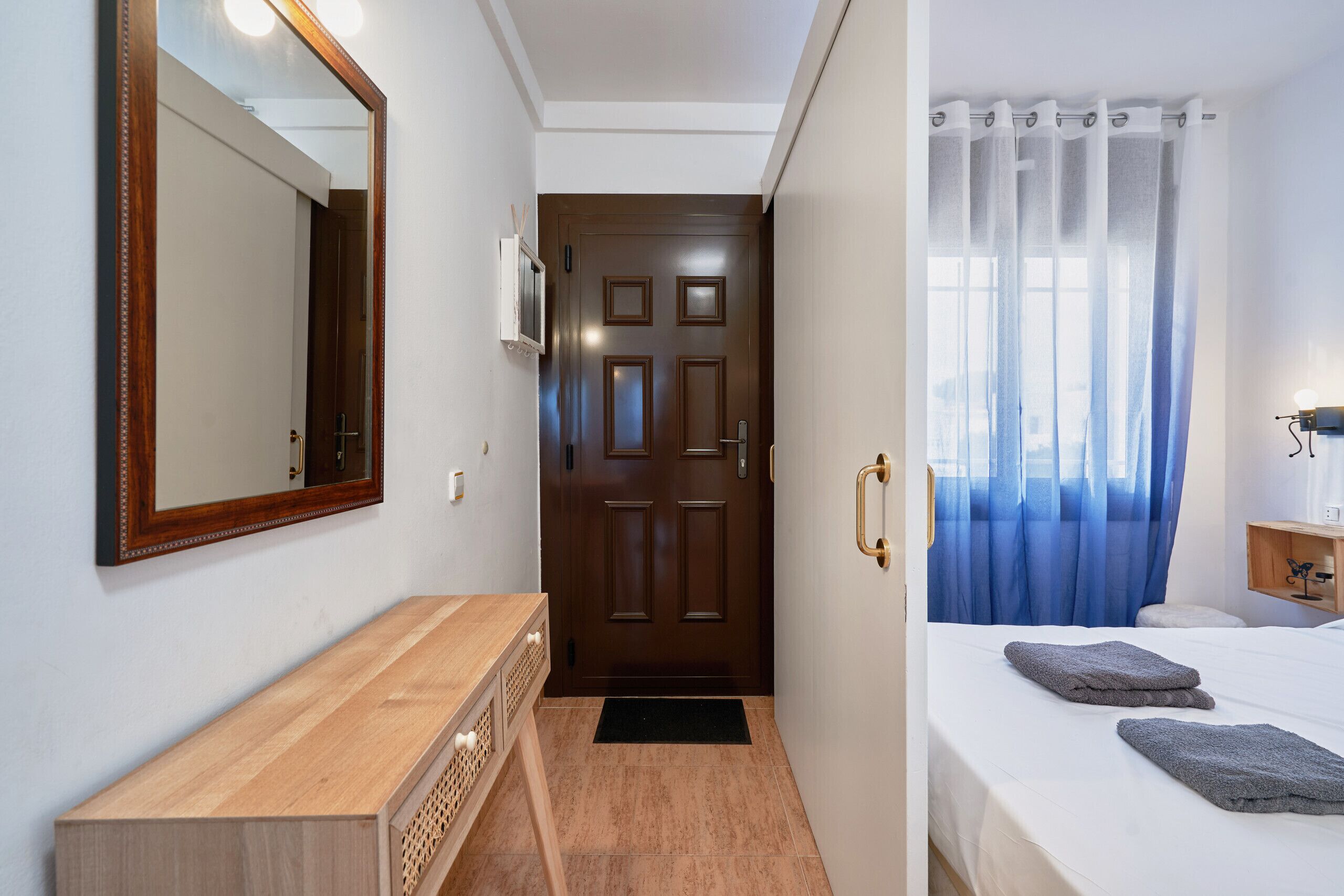 Habitación de hotel con un aparador de madera, una cama con dos toallas, una puerta con espejo y una ventana con cortina azul.