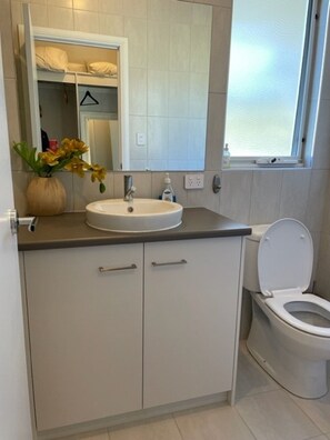 A bathroom with a white sink, a toilet, and a mirror above the sink. There is a window above the toilet and a vase with flowers on the sink counter.
