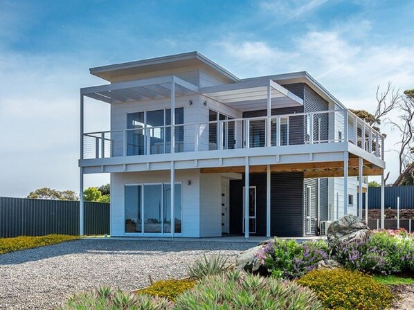 A modern two-story house with a balcony, large windows, and a gravel driveway.