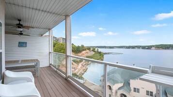 A balcony with a view of a river, a table, and two white chairs.
