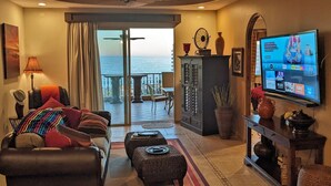 Ein Wohnzimmer mit einer Couch, einem Sessel und einem Fernseher. Durch die Glastüren ist der Blick aufs Meer.