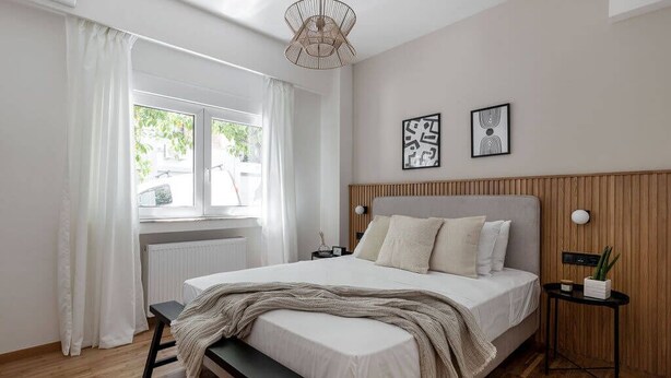 A bedroom with a bed, a bench, a bedside table, a wall-mounted lamp, and a window with curtains. - Dreamy 1BR Apartment in Kolonaki (Athens)