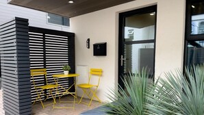 A modern outdoor seating area with yellow chairs and a table, a planter with green plants, and a black mailbox on the wall. - Green Mews 1 By My Getaways (Hove)