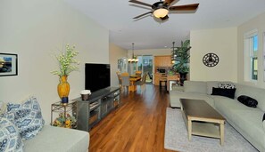 A modern living room with a flat-screen TV, a ceiling fan, a dining area with a table and chairs, and a kitchen in the background.